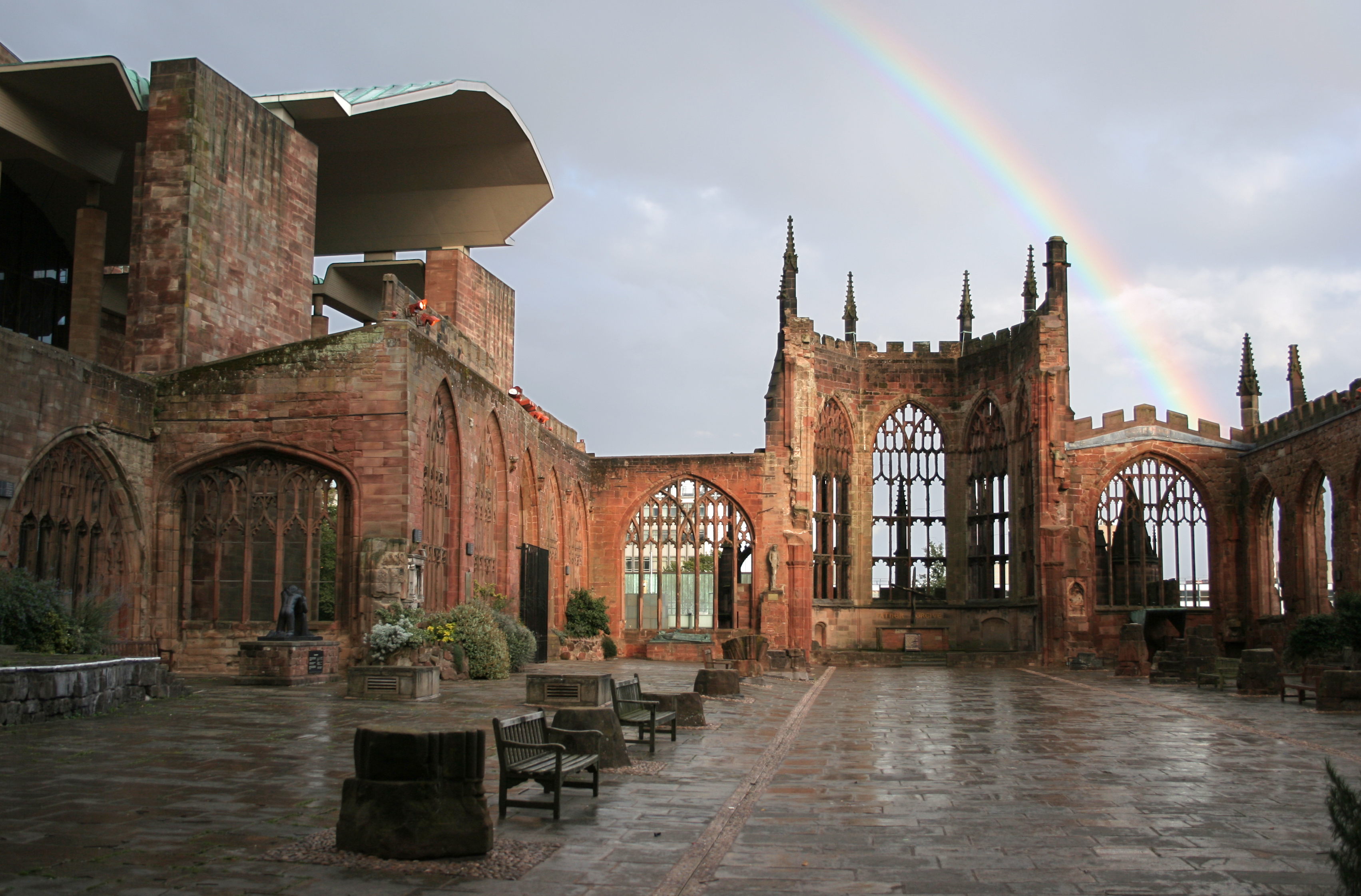A rainbow arches over the open-air ruins of Coventry Cathedral, with stone walls and window tracery surrounding a paved courtyard.