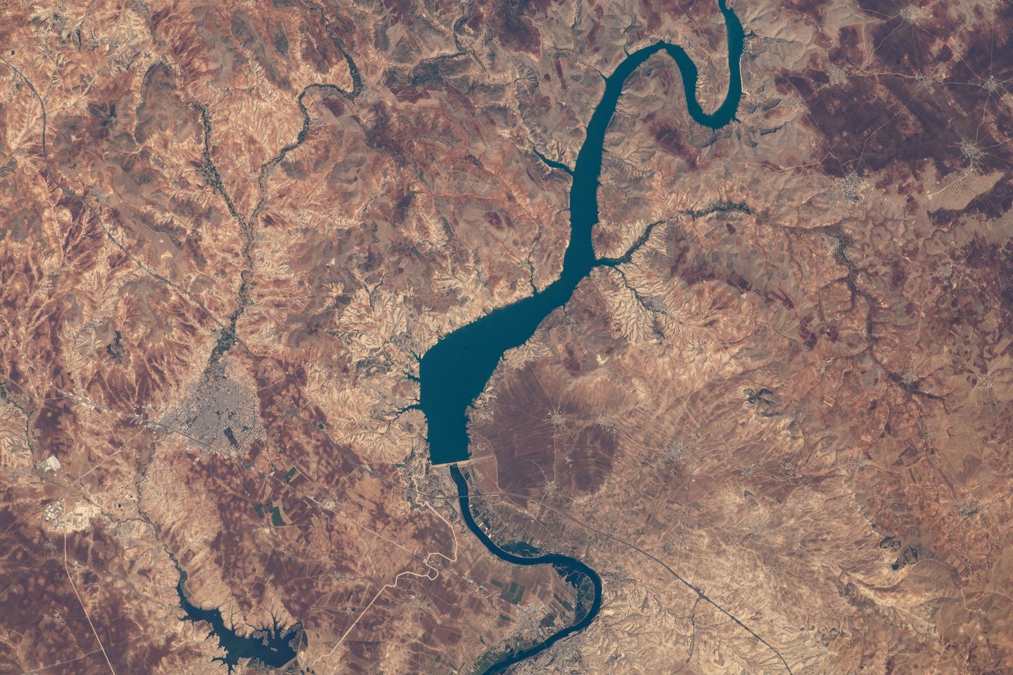 A blue-green river winds through an arid landscape, widening into a reservoir above a dam.
