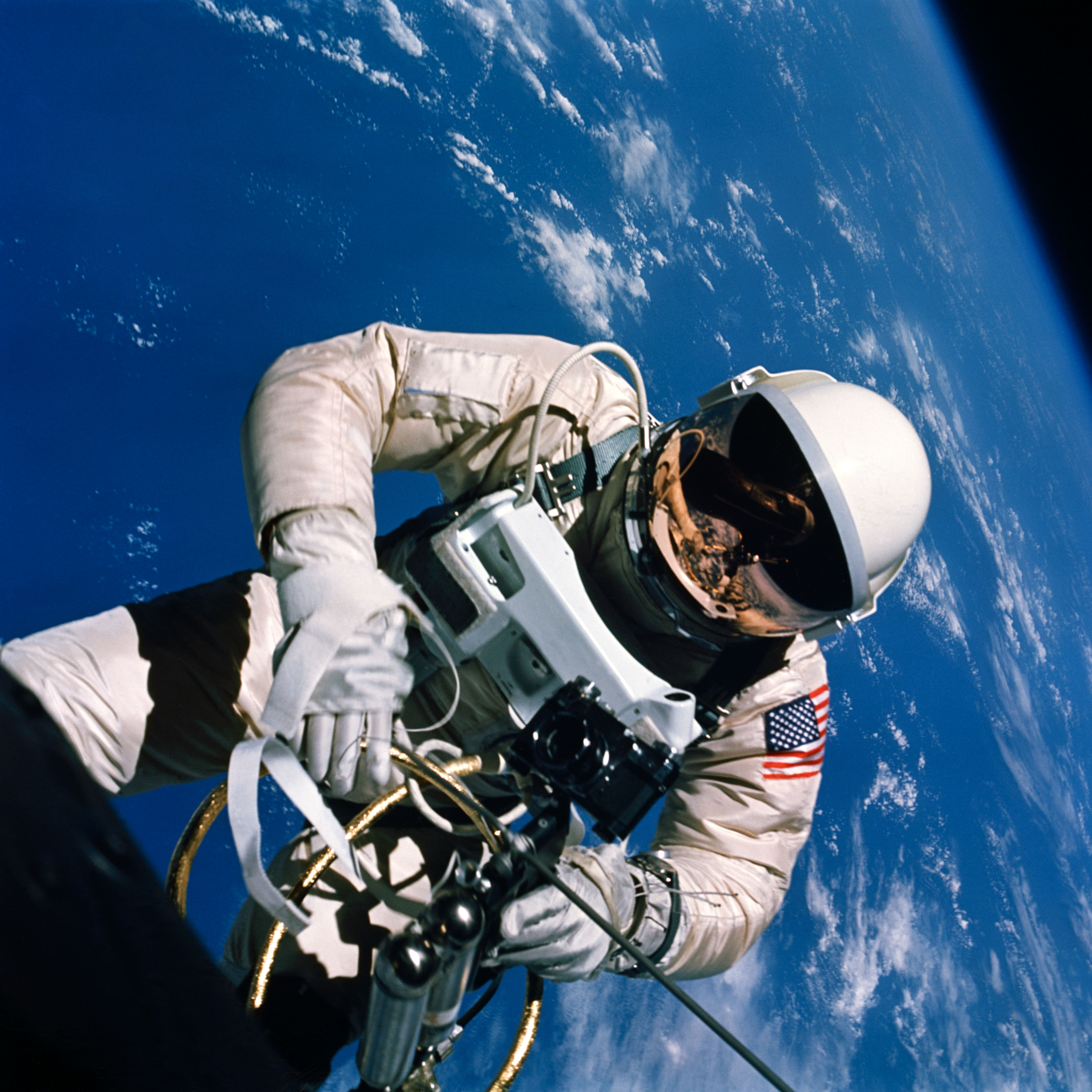 An astronaut in a white suit and reflective visor floating above the blue Earth, holding a maneuvering device.