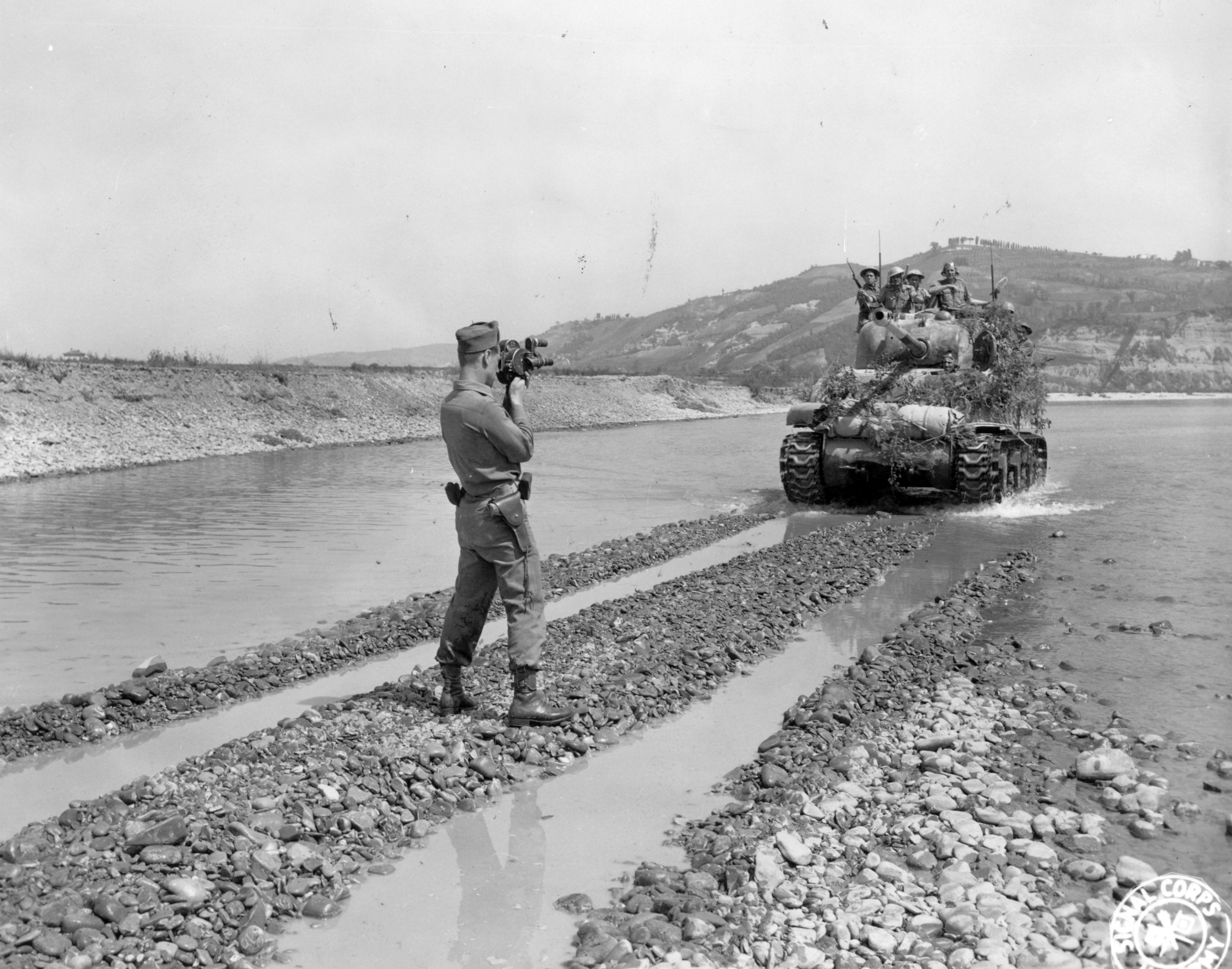A military cameraman films a tank driving across a low river crossing.