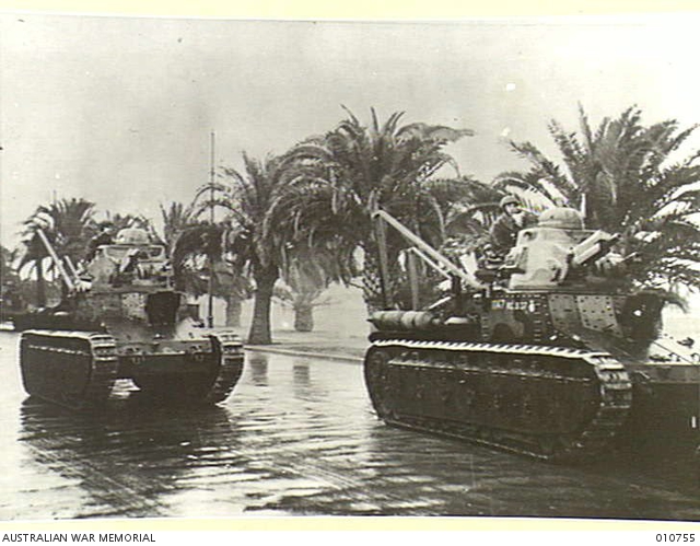 French tanks move along a palm-lined street, with people riding on the nearest tank.