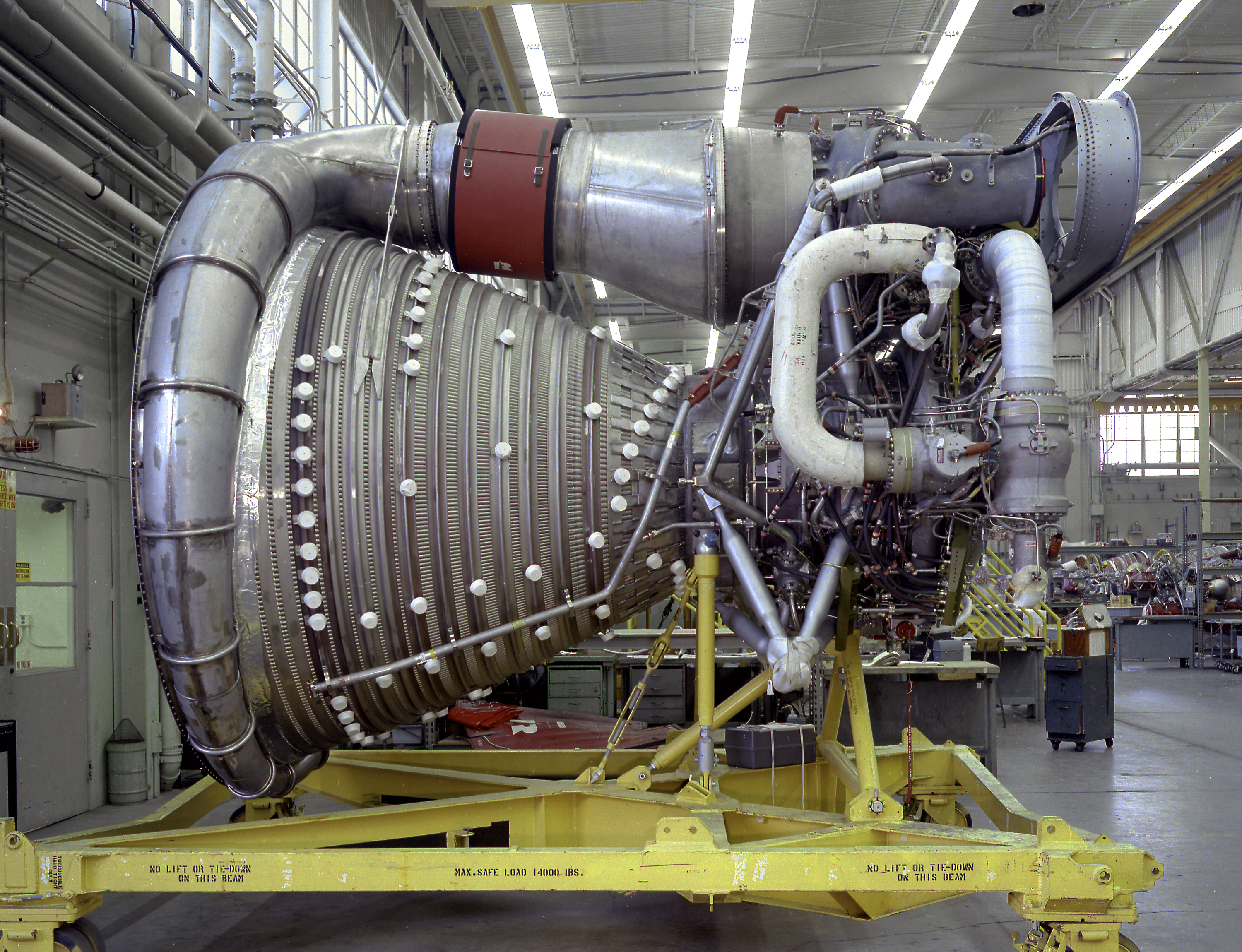 An F-1 engine lying horizontally on a yellow support stand, showing its ribbed nozzle and connected pipework.