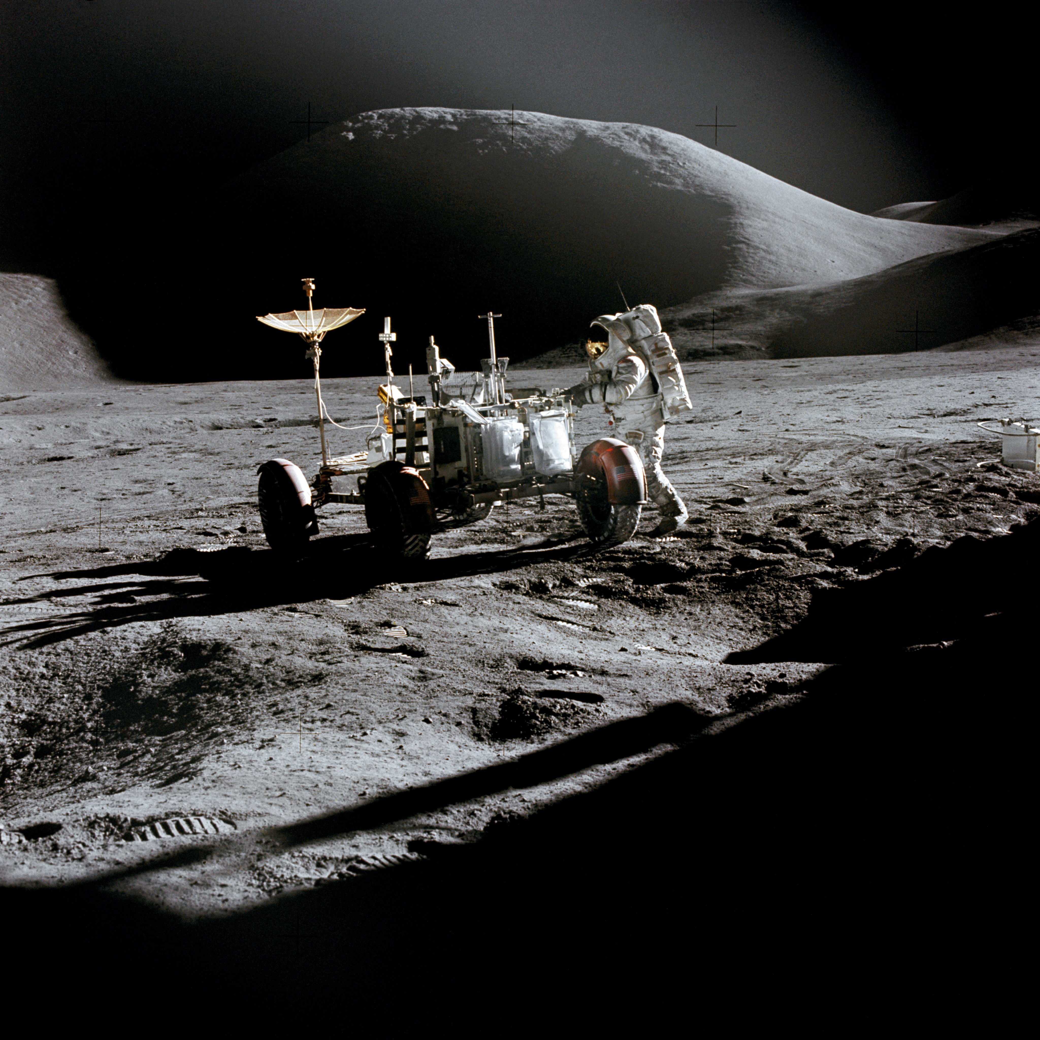 An astronaut standing beside a four-wheeled lunar rover on gray ground below mountain slopes.