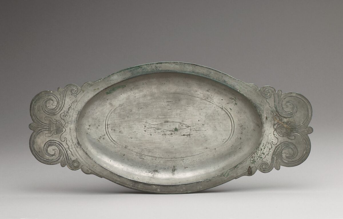 An oval metal platter has a fish engraved in its center and curved decorative handles at both ends.