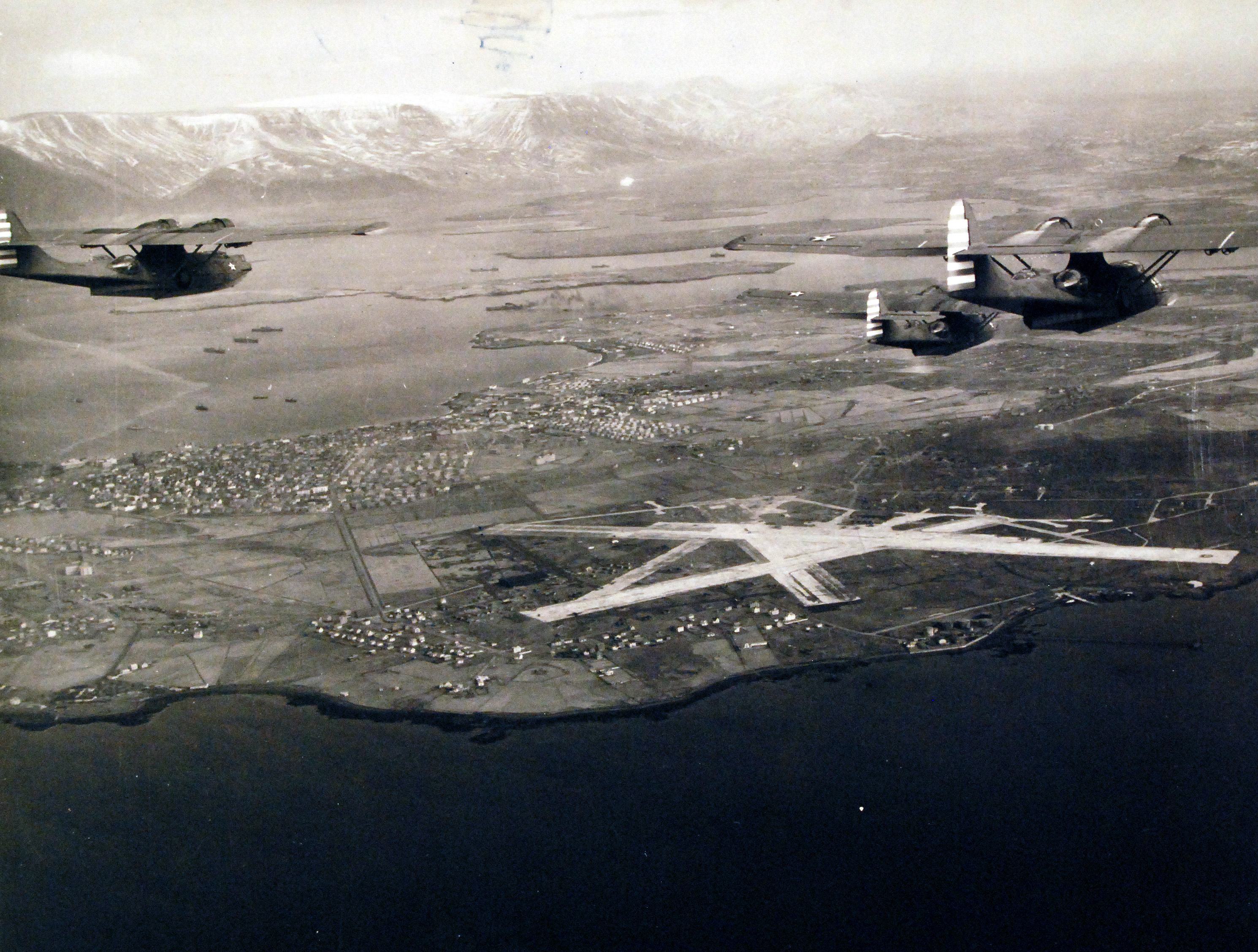 Two Catalina patrol planes fly above Reykjavik’s coastal airfield, with snow-covered hills in the distance.