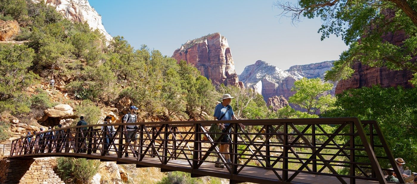 A group of hikers cross a bridge over a river. In the background, Angels Landing stands in view.